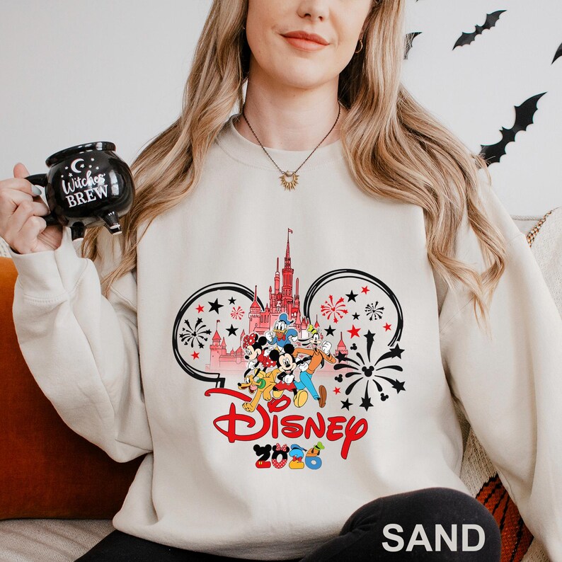 A woman wearing a white sweater with a Disney-themed design, holding a mug with the text "Witch's Brew" on it.