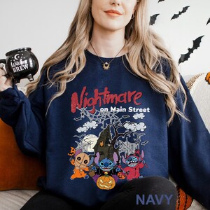 A woman wearing a navy blue sweatshirt with a Halloween-themed design, including characters from the movie "Nightmare on Main Street". She is holding a black mug with a Halloween-themed design.