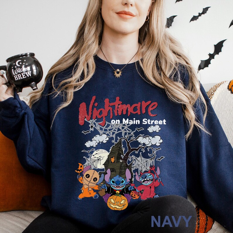 A woman wearing a navy blue sweatshirt with a Halloween-themed design, including characters from the movie "Nightmare on Main Street". She is holding a black mug with a Halloween-themed design.