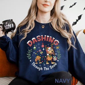 A woman wearing a navy blue sweatshirt with a Christmas-themed design, including a cartoon dog and snowflakes, is holding a mug with the text "DASHING Through The Snow" printed on it.