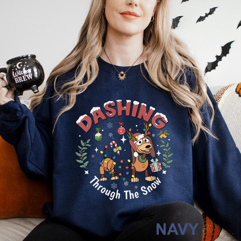A woman wearing a navy blue sweatshirt with a Christmas-themed design, including a cartoon dog and snowflakes, is holding a mug with the text "DASHING Through The Snow" printed on it.
