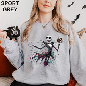 A woman wearing a grey sweatshirt with a graphic of a jack-o-lantern character from the movie " jack-o-lantern". She is holding a black mug with the text "I love coffee" printed on it.
