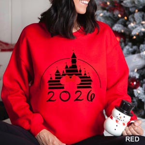 A woman wearing a red sweater with a black silhouette of a castle and the year "2026" printed on it, holding a snowman toy.