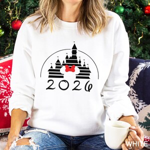 A woman wearing a white sweater with a graphic of a castle and the year "2026" printed on it, sitting in front of a Christmas tree.