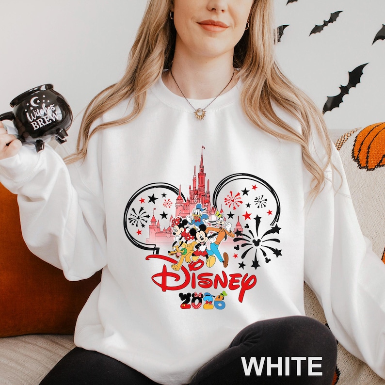 A woman wearing a white sweater with a Disney-themed design, including a castle and fireworks, is holding a mug with the text "Disney 2020" printed on it.