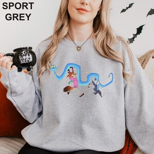 A woman wearing a grey sweatshirt with a cartoon character design is holding a black mug with the text "Cup Brew" printed on it.