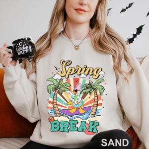 A woman wearing a white sweatshirt with a colorful graphic design of a cartoon character surfing on a wave, holding a mug with the text "Witch's Brew" printed on it.
