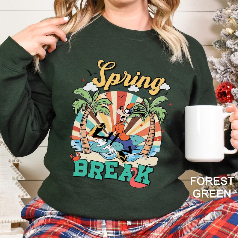 A woman wearing a green sweatshirt with a graphic design of a cartoon character and the text "Spring Break" is holding a white mug.