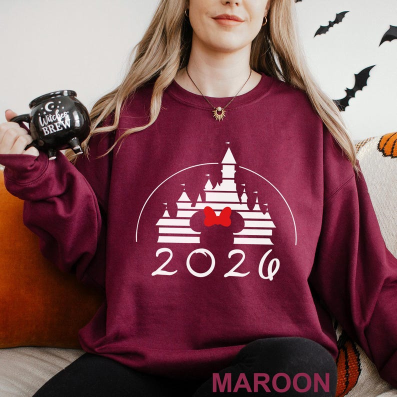 A woman wearing a maroon sweater with a white castle graphic and the year "2020" printed on it, holding a black mug with the text "C witch brew" printed on it.