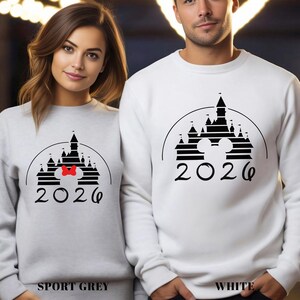 A woman and a man standing side by side wearing matching white sweatshirts with a black and white castle graphic and the year "2026" printed on them.