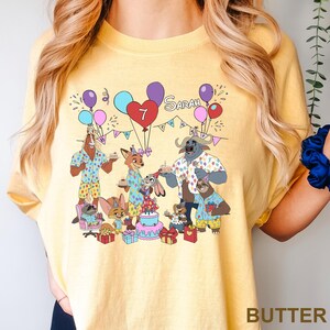 A yellow t-shirt with a cartoon illustration of a group of animals, including a dog, cat, and cow, celebrating a birthday.