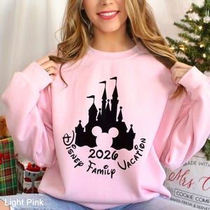 A woman wearing a pink sweatshirt with a Disney castle graphic and text, smiling and posing in front of a Christmas tree.