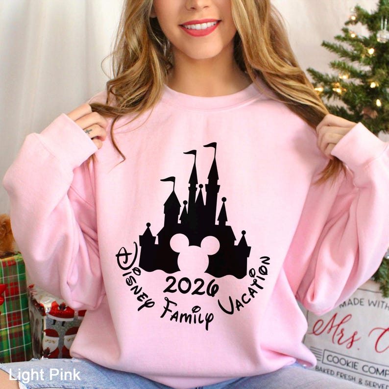 A woman wearing a pink sweatshirt with a Disney castle graphic and text, smiling and posing in front of a Christmas tree.