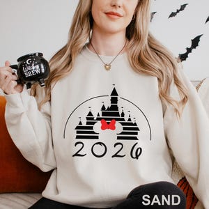 A woman wearing a white sweater with a black and white castle graphic and the year "2026" printed on it, holding a mug with the text "Witch's Brew" printed on it.
