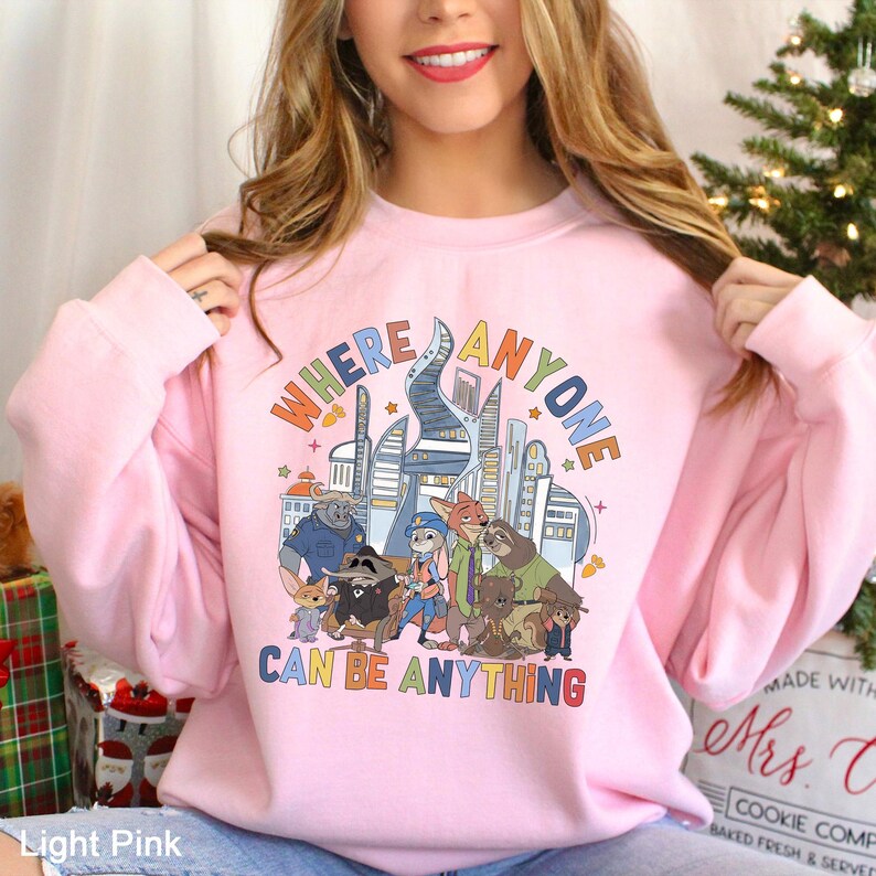 A woman wearing a pink sweatshirt with a graphic design featuring cartoon characters and the text "WHERE ANYONE CAN BE ANYTHING".