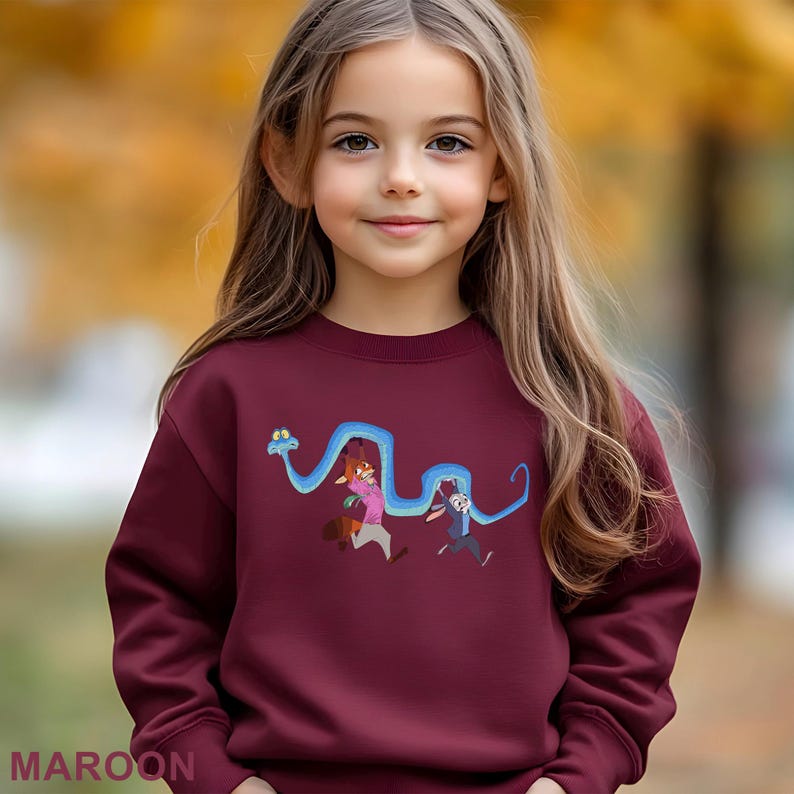 A young girl with long blonde hair is wearing a maroon sweater with a cartoon character design on it.