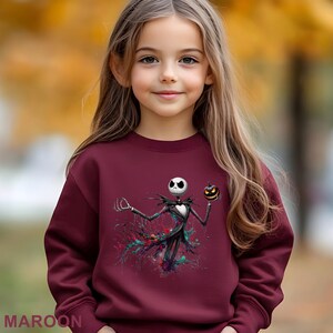 A young girl wearing a maroon sweater with a graphic of a skeleton figure holding a jack-o-lantern.