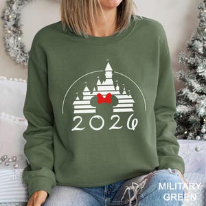 A woman wearing a green sweatshirt with a white castle graphic and the year "2026" printed on it, sitting in front of a Christmas tree.
