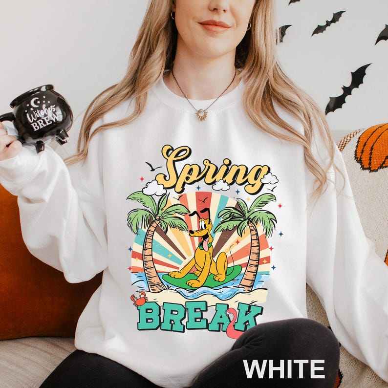 A woman wearing a white sweatshirt with a cartoon image of a dog surfing on a beach, holding a mug with the text "Spring Break" printed on it.