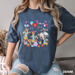 A person wearing a blue t-shirt with a graphic design depicting a birthday party scene with various cartoon animals and decorations.