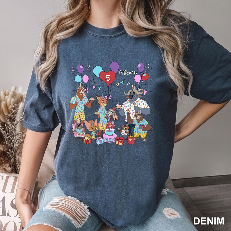 A person wearing a blue t-shirt with a graphic design depicting a birthday party scene with various cartoon animals and decorations.