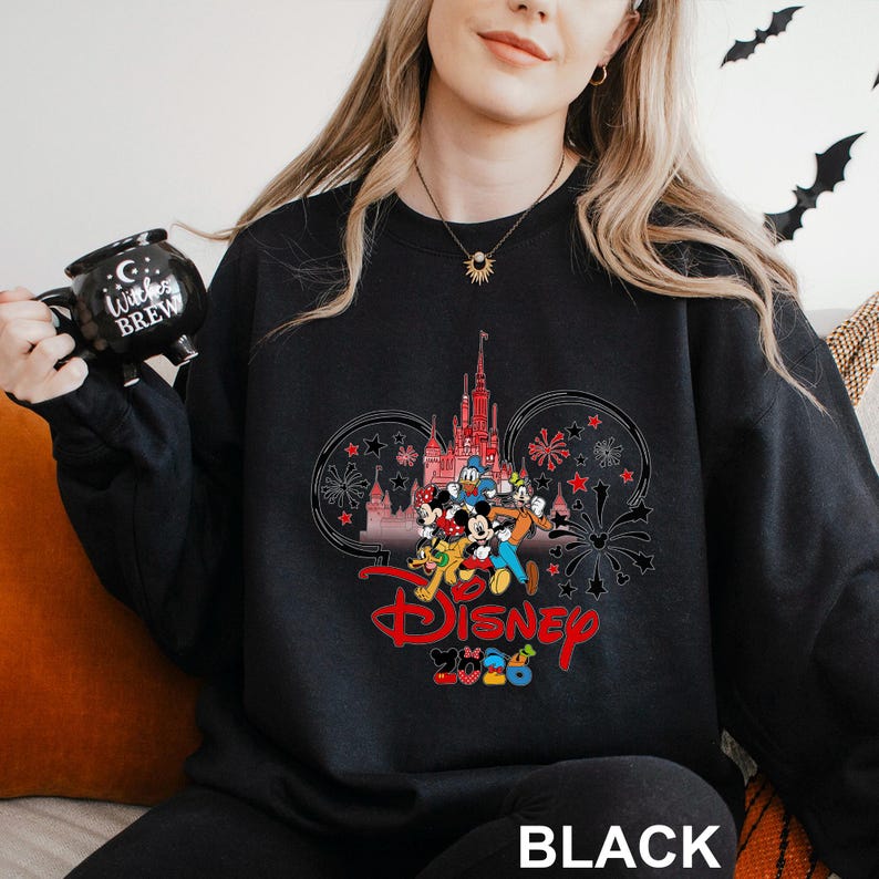 A woman wearing a black sweatshirt with a Disney-themed design, including Mickey Mouse and other Disney characters, is holding a black mug with the text "Disney BREW" printed on it.