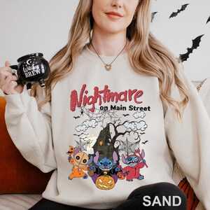A woman wearing a white sweatshirt with a Halloween-themed design, including a spooky house, bats, and a character from the movie "Nightmare on Main Street". She is holding a mug with the text "Witch's Brew" printed on it.