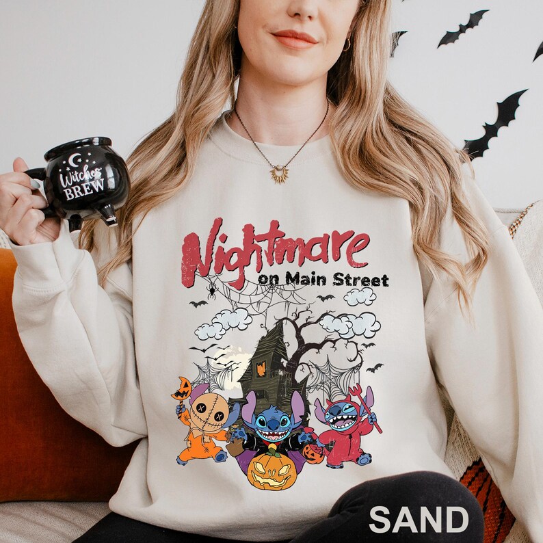 A woman wearing a white sweatshirt with a Halloween-themed design, including a spooky house, bats, and a character from the movie "Nightmare on Main Street". She is holding a mug with the text "Witch's Brew" printed on it.