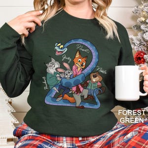 A woman wearing a green sweater with a cartoon illustration of various animals, including a snake, rabbit, and fox, along with the text "Nate", "Jude", and "Nelly". She is holding a white mug.