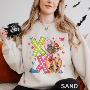 A woman wearing a white sweatshirt with a graphic of cartoon animals and a large "X" on it, holding a mug with the text "Witch's Brew" printed on it.
