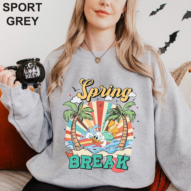 A woman wearing a grey sweatshirt with a colorful graphic design depicting a beach scene and the text "Spring Break" printed on it.