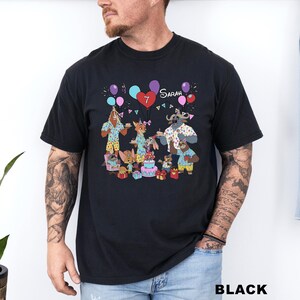 A black t-shirt with a colorful graphic design depicting a group of cartoon animals, including a dog, cat, and cow, celebrating a birthday with balloons and presents.