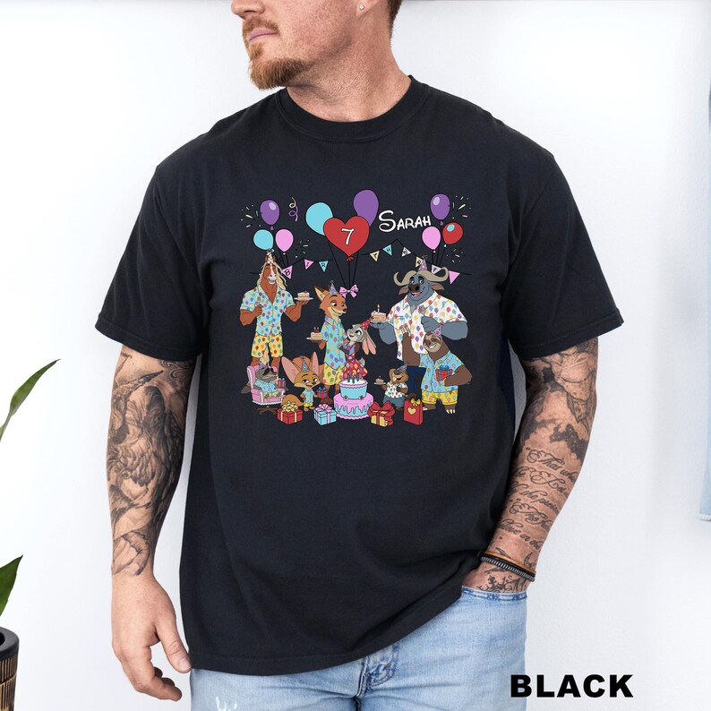 A black t-shirt with a colorful graphic design depicting a group of cartoon animals, including a dog, cat, and cow, celebrating a birthday with balloons and presents.