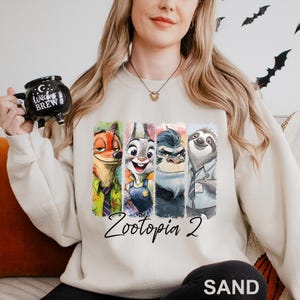 A woman wearing a white sweatshirt with a graphic of four cartoon animal characters from the movie "Zootopia 2" and holding a mug with the text "Witch's Brew" printed on it.