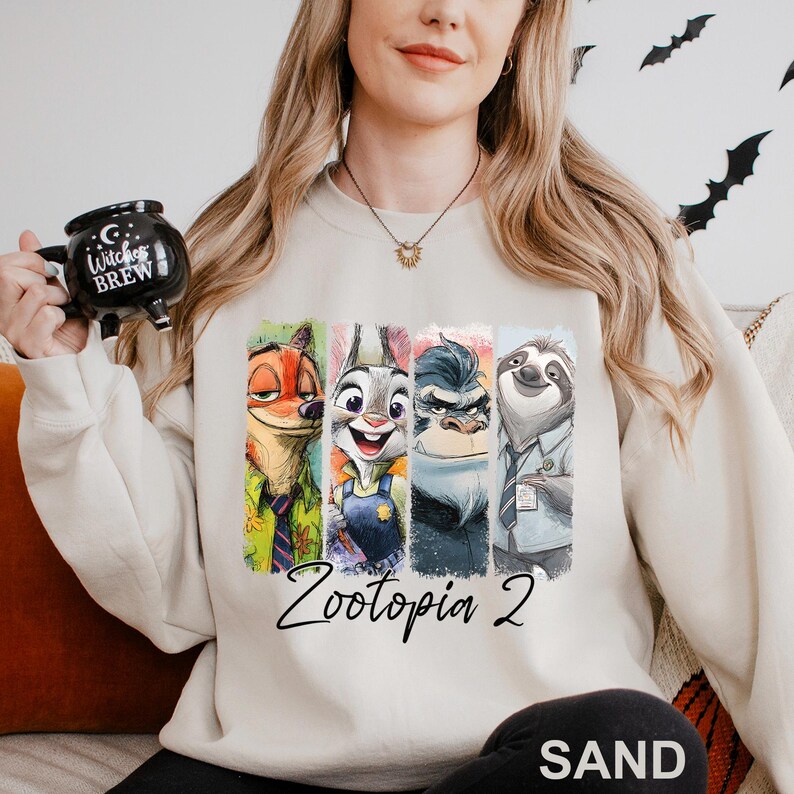 A woman wearing a white sweatshirt with a graphic of four cartoon animal characters from the movie "Zootopia 2" and holding a mug with the text "Witch's Brew" printed on it.