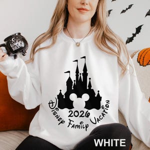 A woman wearing a white sweater with a black silhouette of a castle and the text "Disney Family Vacation" printed on it, holding a mug with a black design.