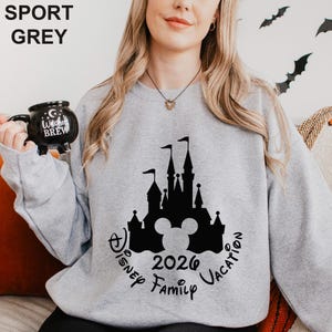 A woman wearing a grey sweatshirt with a black silhouette of a castle and Mickey Mouse, holding a black mug with the text "Disney BREW" on it.