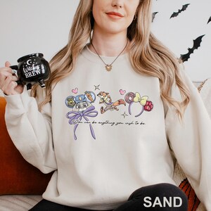 A woman wearing a white sweatshirt with a graphic design of cartoon characters and text, holding a black mug with a witch's brew design.