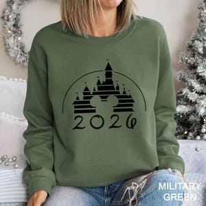 A woman wearing a green sweatshirt with a graphic of a castle and the year "2026" printed on it.