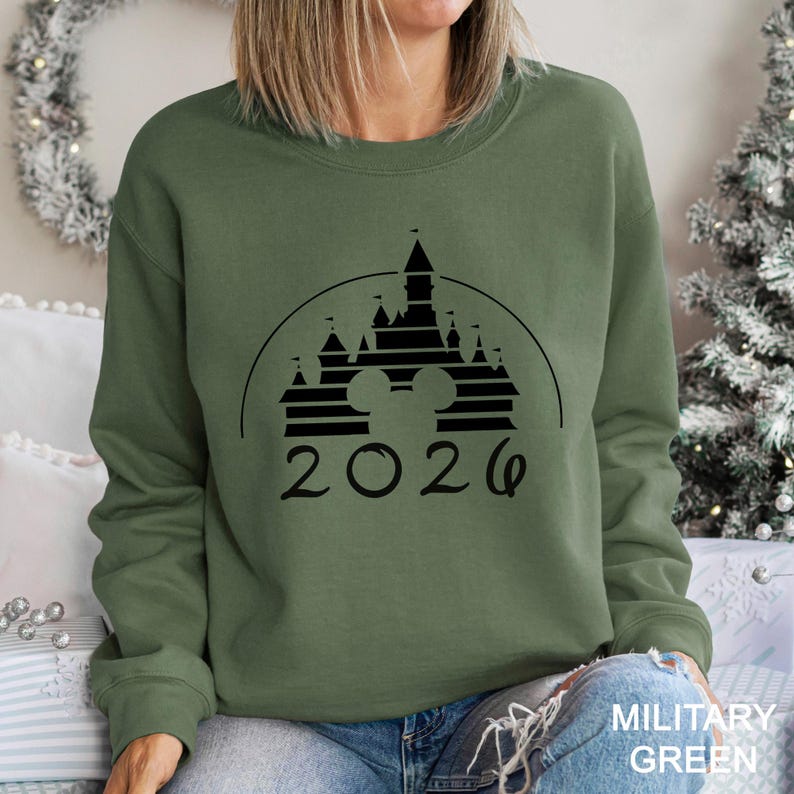 A woman wearing a green sweatshirt with a graphic of a castle and the year "2026" printed on it.