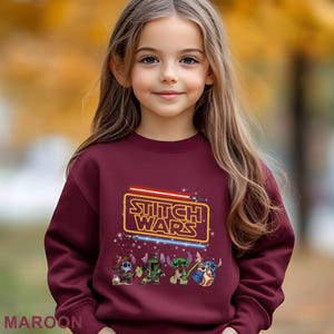 A young girl wearing a maroon sweater with a Star Wars logo and characters printed on it.
