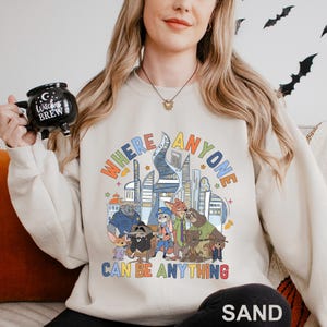 A woman wearing a white sweatshirt with a colorful graphic design and text, holding a black mug with a whimsical design.