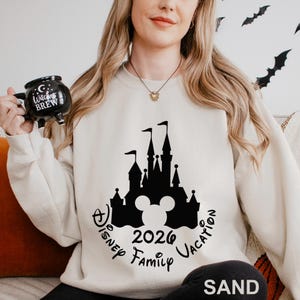 A woman wearing a white sweater with a black castle silhouette and text on it, holding a mug with a black design.