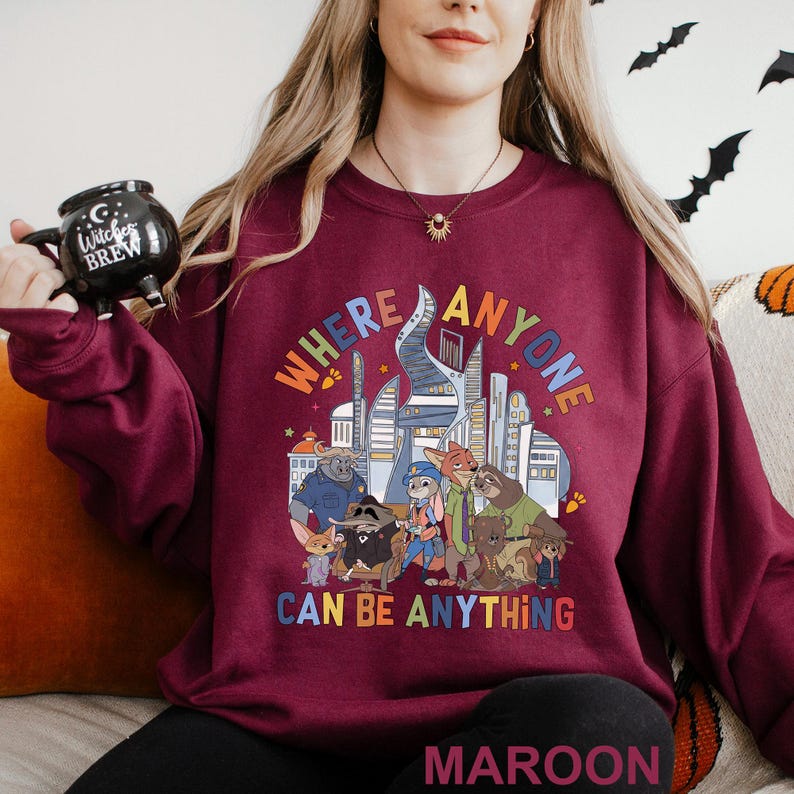 A woman wearing a maroon sweatshirt with a graphic design of cartoon animals and the text "WHERE ANYONE CAN BE ANYTHING" is sitting on a couch.