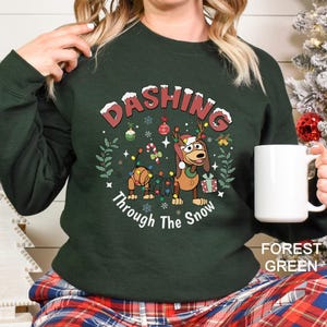 A woman wearing a green sweater with a Christmas-themed design, including a cartoon dog and snowflakes, holding a white mug.