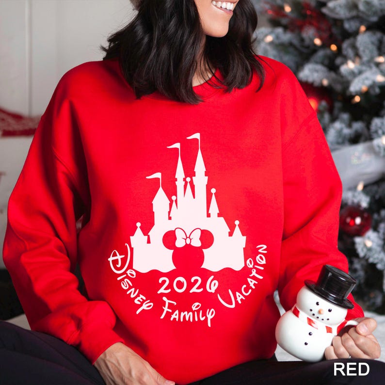 A woman wearing a red sweater with a white castle and Minnie Mouse design, holding a snowman toy, and smiling.