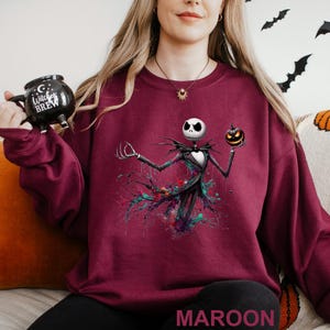 A woman wearing a maroon sweater with a graphic of a jack-o-lantern and a mug with the text "C witch brew" printed on it.