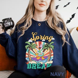 A woman wearing a navy blue sweatshirt with a colorful graphic design of a cartoon character surfing on a wave, surrounded by palm trees and a sunburst pattern.