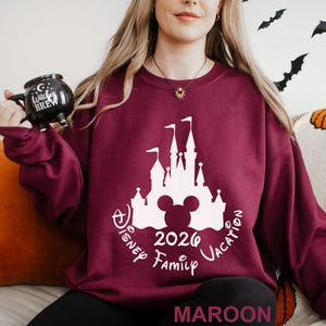 A woman wearing a maroon sweater with a white castle and Mickey Mouse design, holding a black mug with the text "Disney Family Vacation" printed on it.