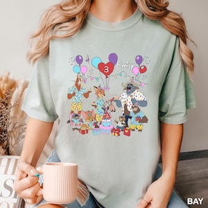 A person is wearing a t-shirt with a cartoon illustration of a birthday party scene featuring animals and a cake.